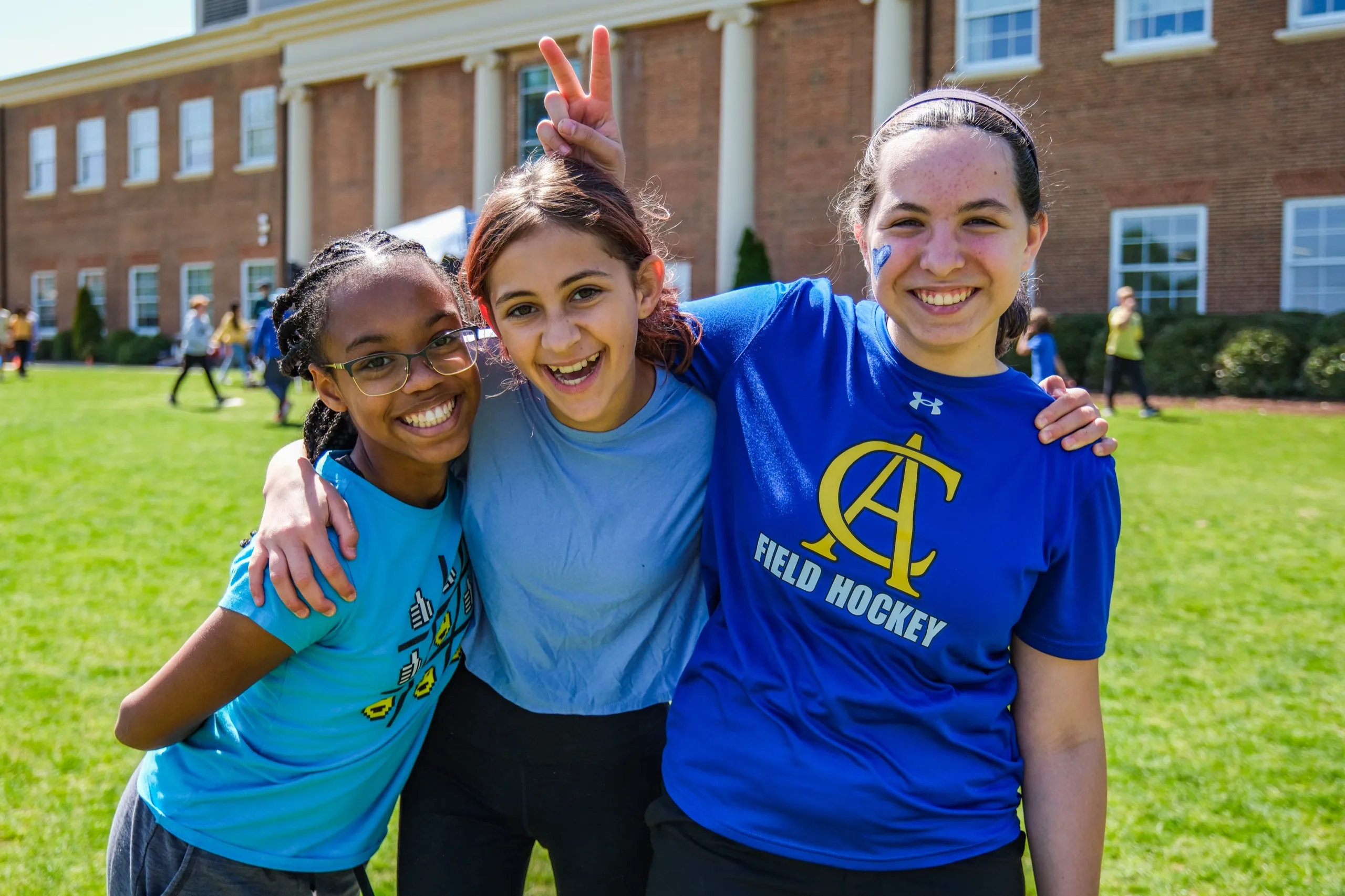 Three students in Cary Academy shirts hug and smile on the lawn in front of the main building