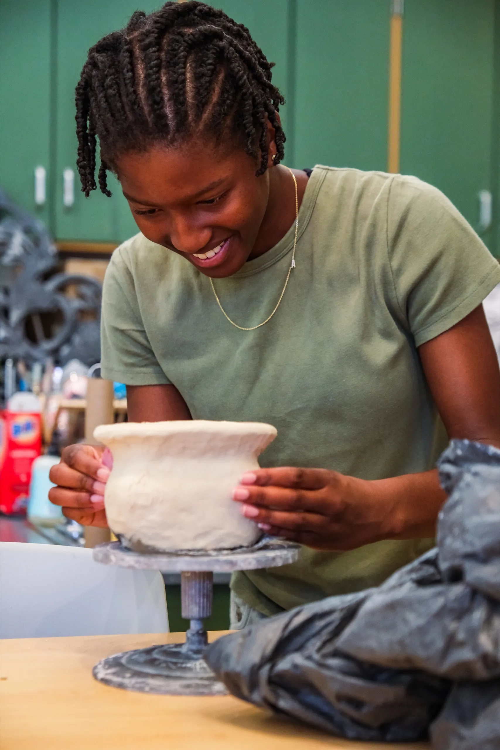 A student smiles as she shapes a tall clay vase on the wheel