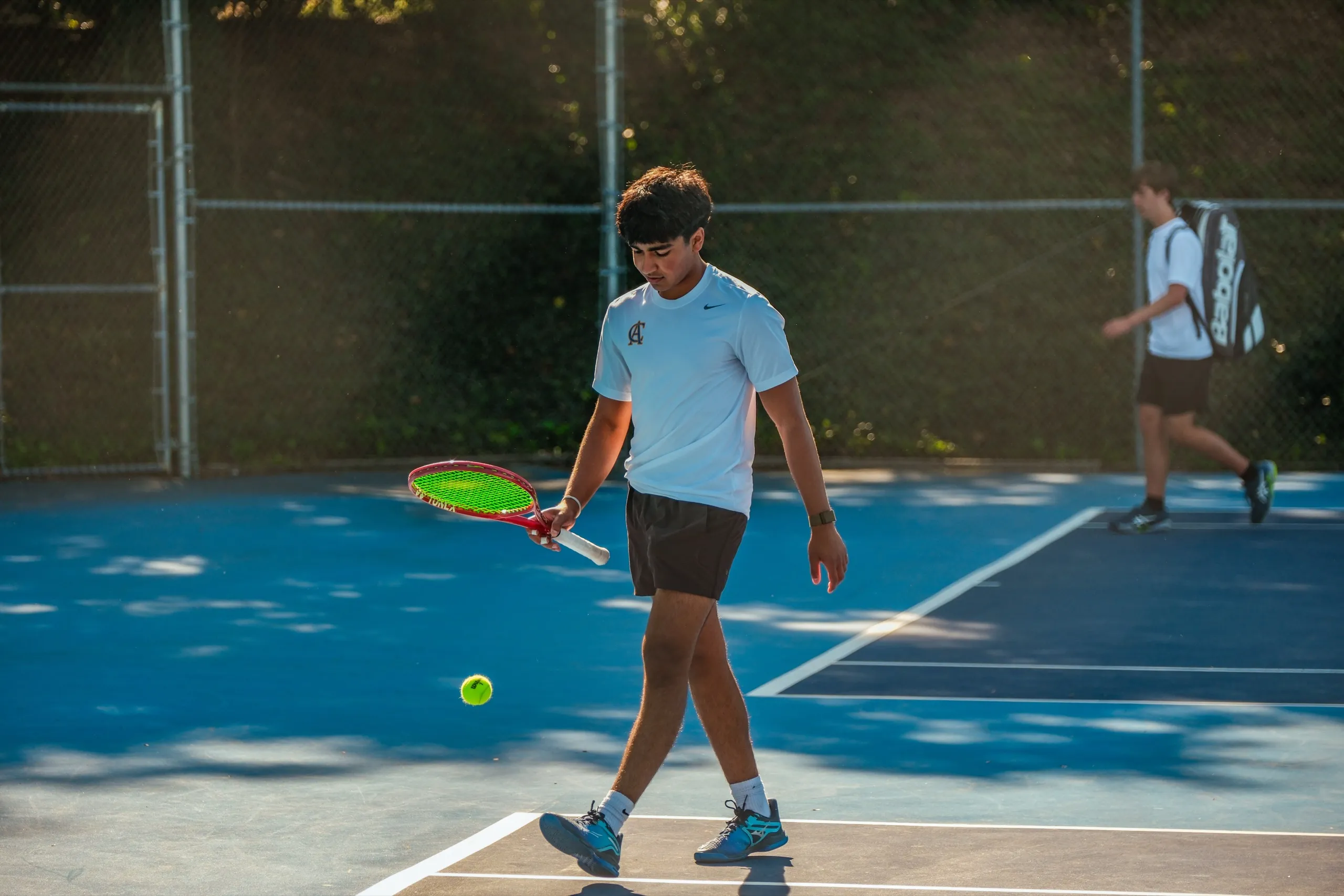 An Upper School student walks off the tennis court holding a racket, a ball bouncing beside him